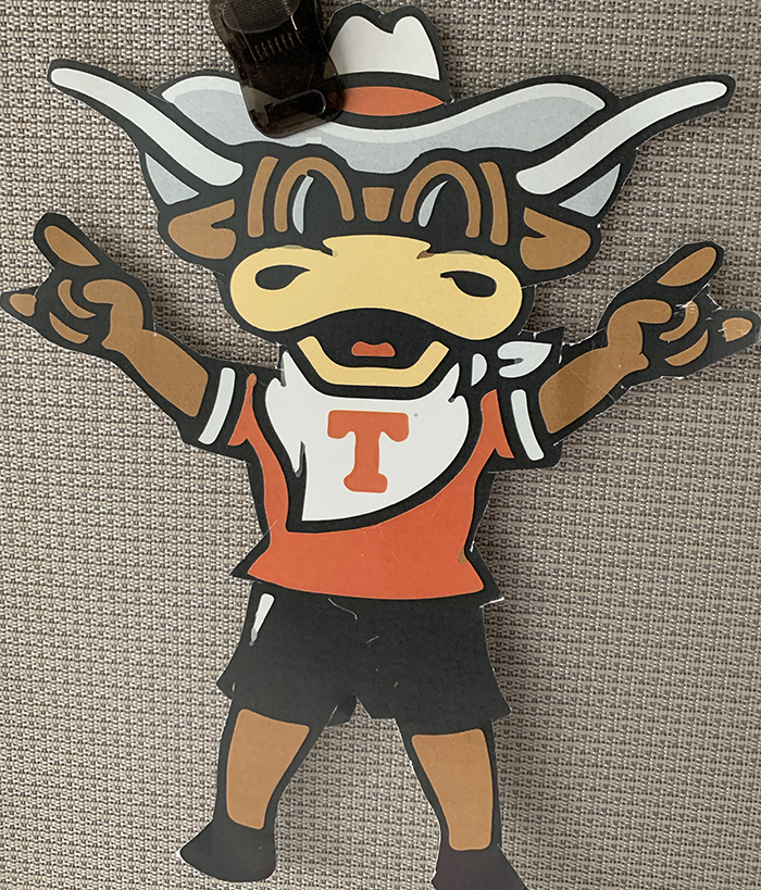 An image of a piece of team paraphernalia from the University of Texas Longhorns. A cartoon representation of the UT mascot wears a cowboy hat and flashes the "Hook 'em Horns" sign with his suspiciously human-looking hands. 
